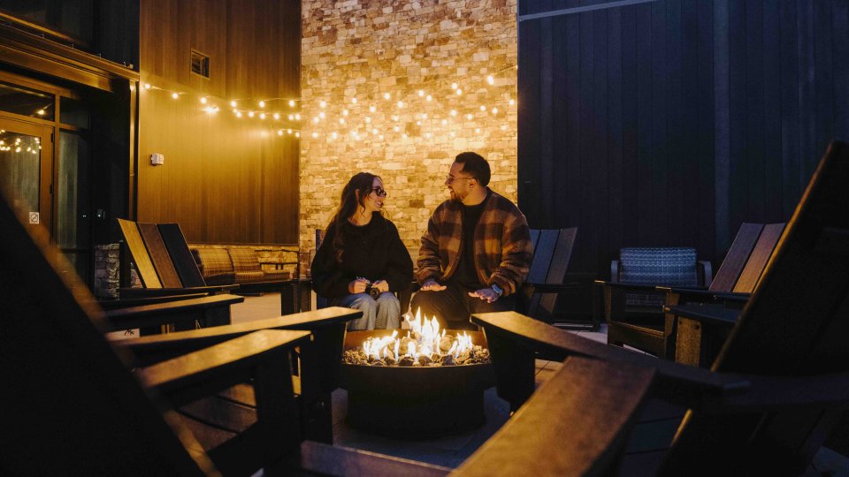 A couple sits by a glowing fire pit, surrounded by wooden chairs. String lights hang above, creating a cozy, intimate atmosphere on a patio at dusk.