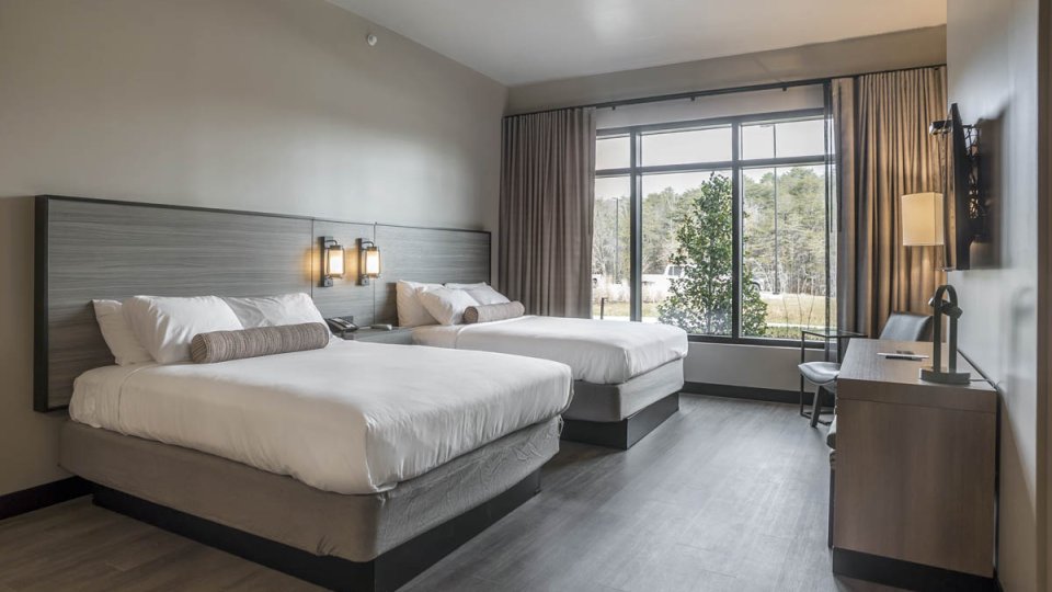 Bedroom with two queen sized beds with modern lighting, a large window, a chair is in the corner, and a television is facing the beds