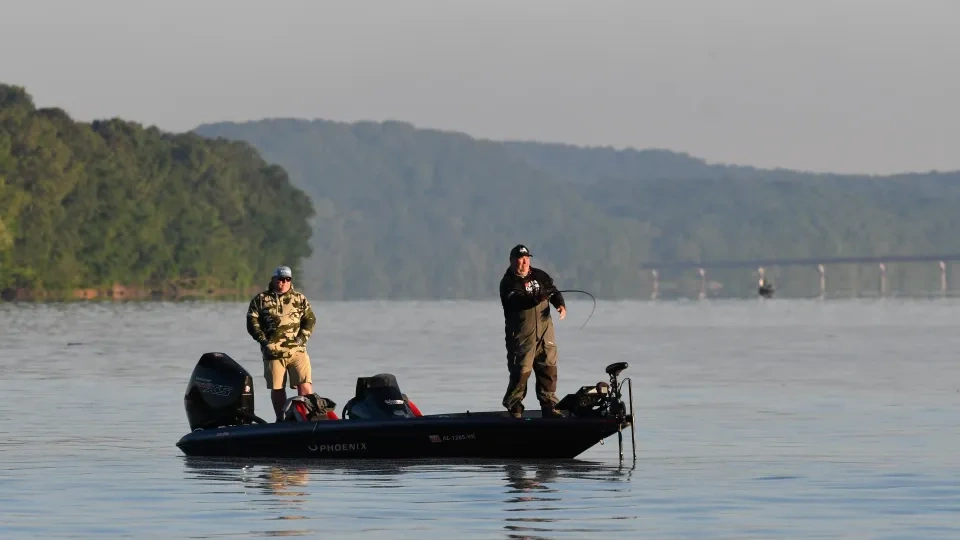 Two people fishing on a boat in a calm lake, surrounded by hills.