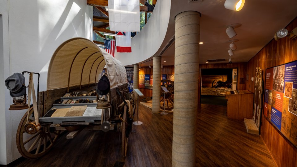 A historical exhibit shows a covered wagon with hats on its side. Flags hang overhead. Informational displays line the wooden walls, creating an educational atmosphere.