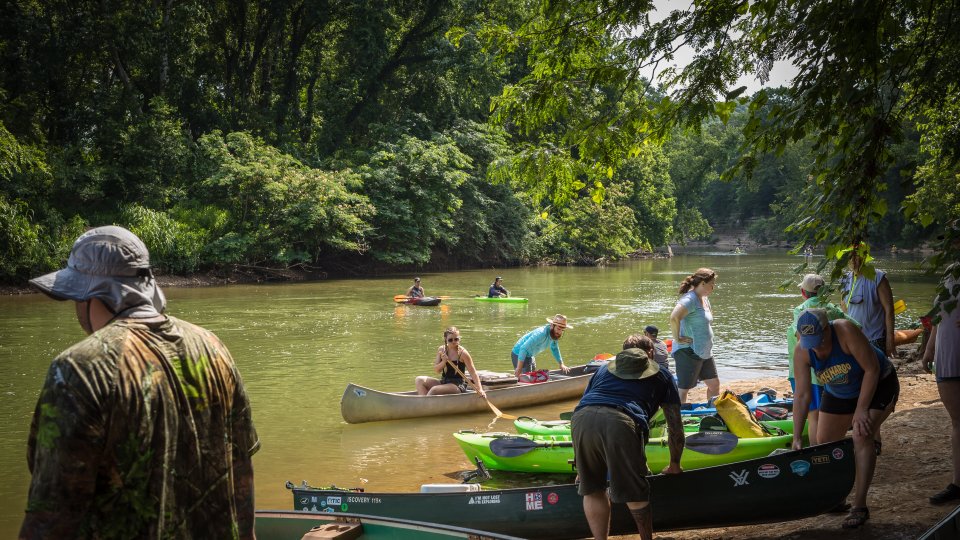 People launch canoes and kayaks into a calm river surrounded by lush green trees, creating a lively and adventurous atmosphere.