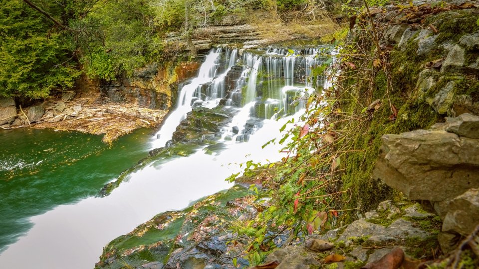 Cascading waterfall flows over tiered rocks into a serene green pool, surrounded by lush forest. A stone ledge with moss and leaves frames the scene.