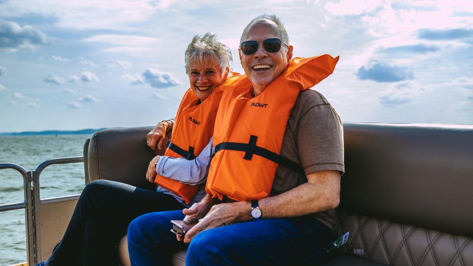 Elderly couple wearing orange life jackets sit smiling on a boat, with a calm lake and cloudy sky in the background. They appear joyful and relaxed.