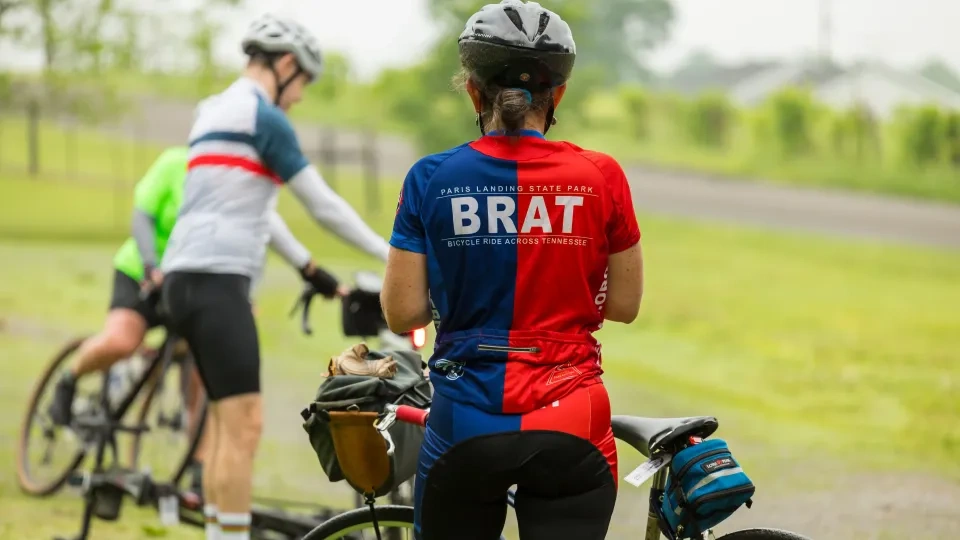 Cyclists in colorful jerseys resting with bicycles on a grassy path.