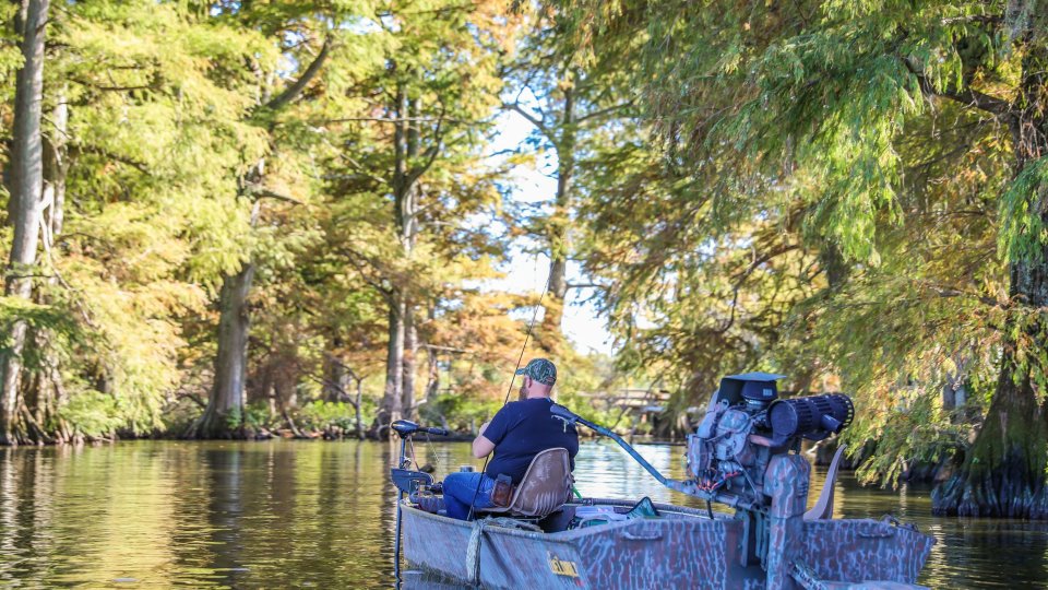 A man in a blue shirt sits in a small motorboat, fishing on a calm river surrounded by tall, lush trees tinged with autumn colors, evoking a peaceful, serene atmosphere.