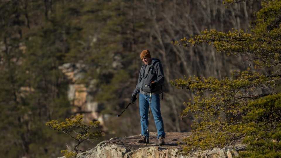 Person standing on a rocky cliff edge, surrounded by trees, wearing a jacket and jeans. The scene is calm, evoking a sense of adventure and solitude.