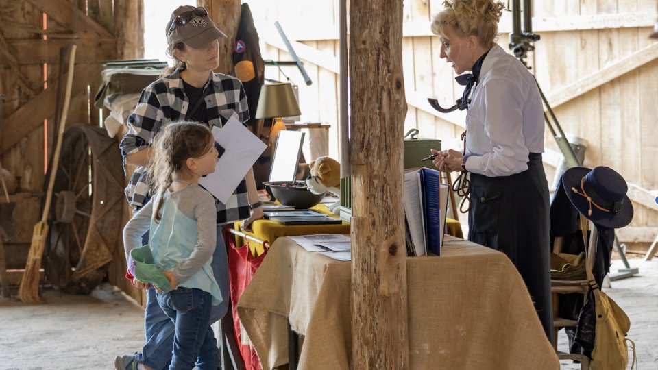 A child and adult visit a historical booth inside a rustic barn. A person in period costume engages them with antique items on a burlap-covered table.
