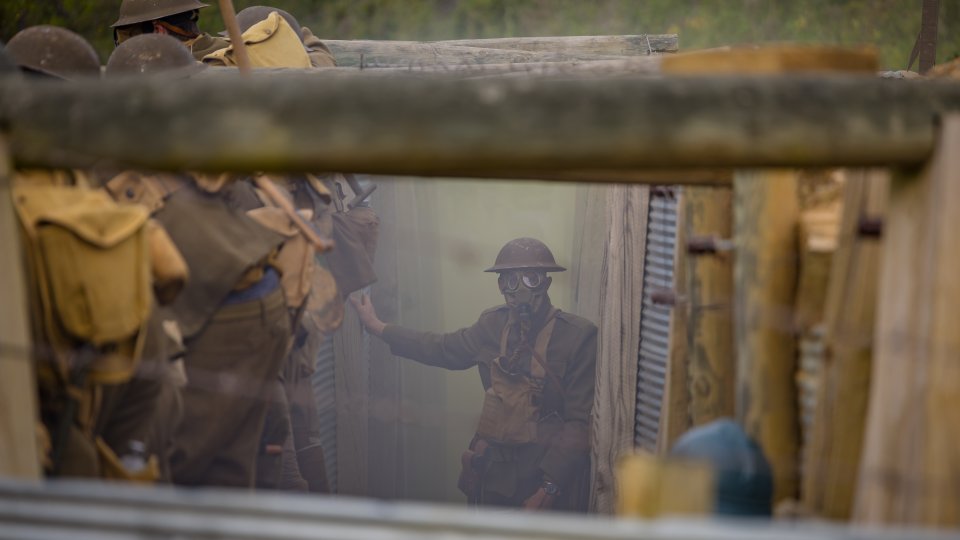 A group of soldiers in World War I uniforms stand in a smoky trench. The focus is on one soldier wearing a gas mask, conveying a tense atmosphere.