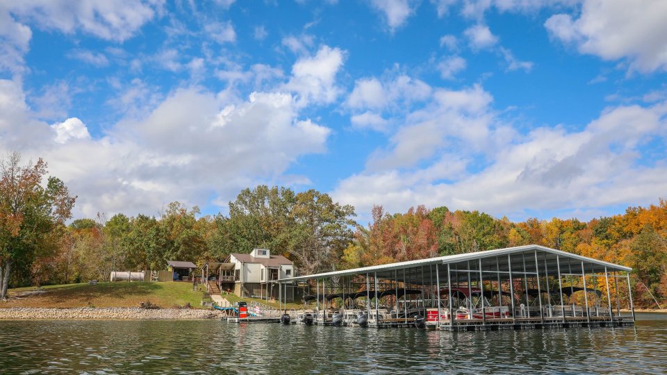 Lakeside view with a house surrounded by autumn trees, and a large boathouse on the water under a bright blue sky with fluffy clouds. Calm and scenic.