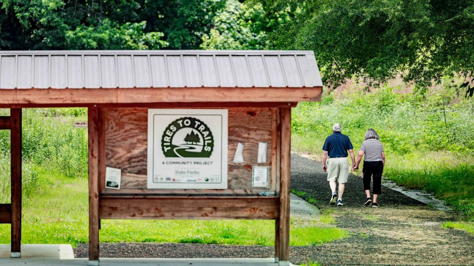 A couple holds hands, walking on a trail near a wooden kiosk with a "Tires to Trails" community project sign. The scene conveys a peaceful, nature-filled atmosphere.