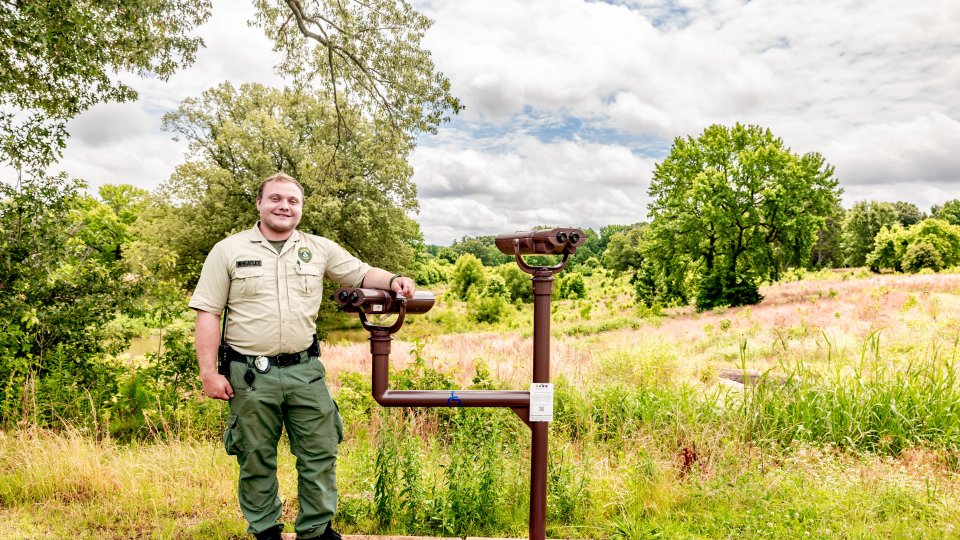 A park ranger stands smiling beside binoculars under a cloudy sky in a lush, green field surrounded by trees, conveying tranquility and nature.