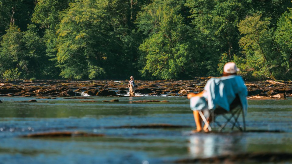 Man sitting in a chair outside by water while a man in the distance is in the water fishing