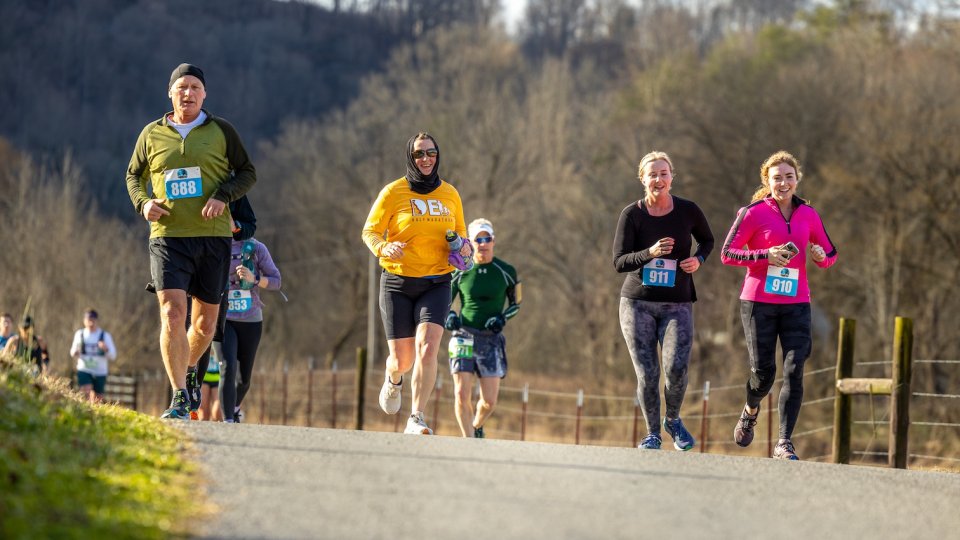 A group of runners on a rural path, wearing colorful athletic gear. The scene is bright and lively, with trees and blue skies in the background.