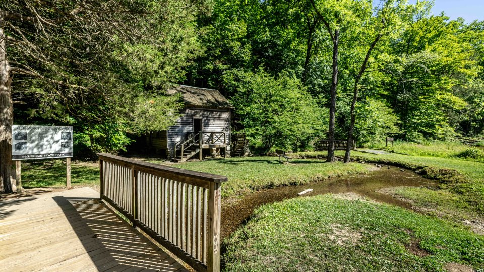 Old wooden cabin by a pond in a lush green forest, viewed from a wooden deck.