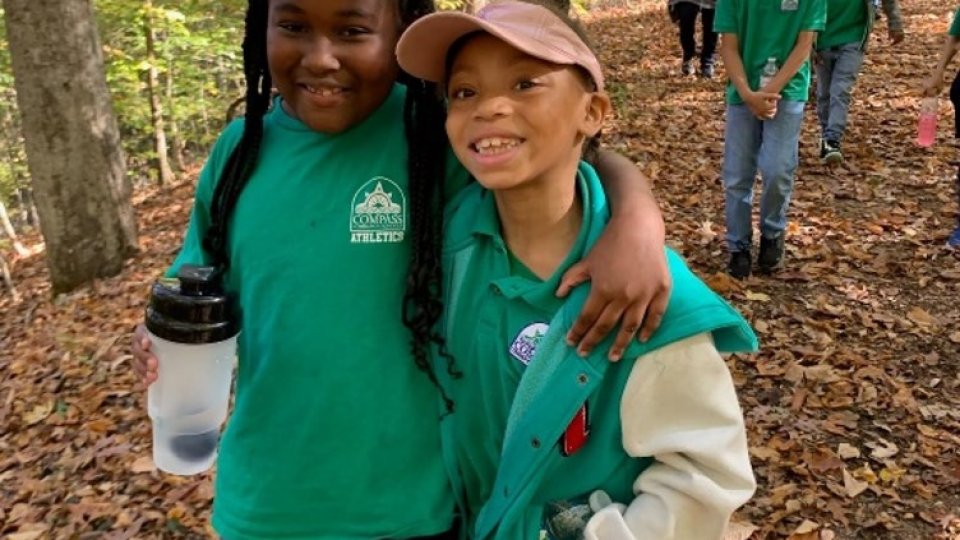 Two girls smiling in a forest, wearing green shirts and carrying water bottles.