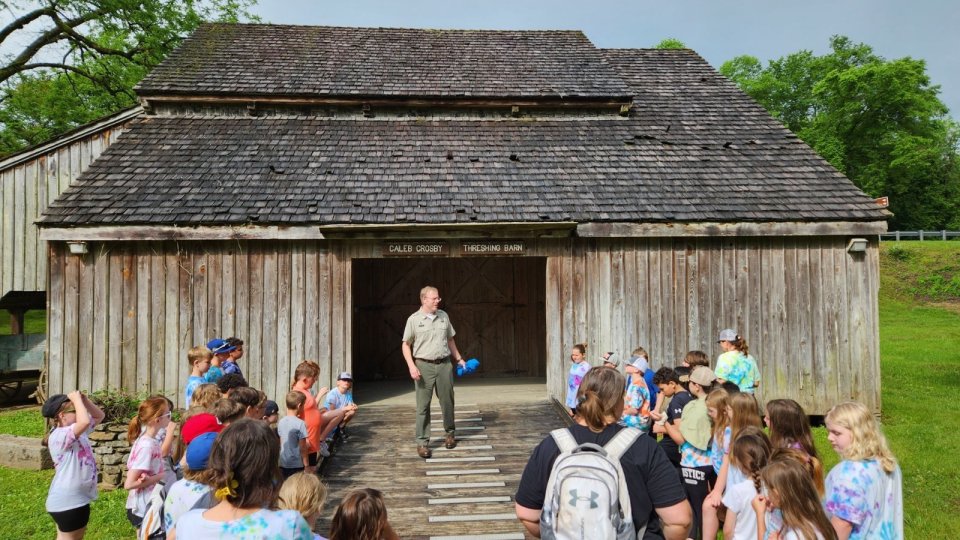 Group of children listening to a guide outside an old wooden barn.