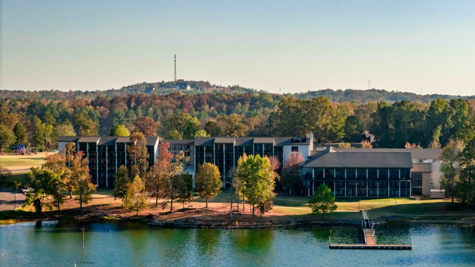 Lakeside resort buildings surrounded by lush trees with a hill in the background.