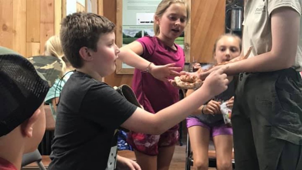 In a nature center, children are interacting with a masked guide holding a snake. Taxidermy birds adorn the wooden walls, creating an educational atmosphere.