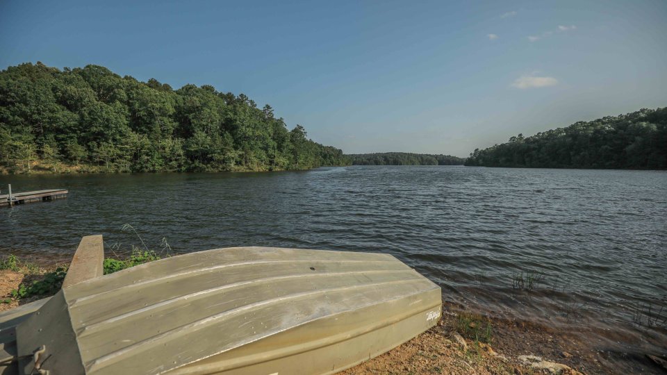 Overturned boat on shore beside a calm lake, surrounded by trees.