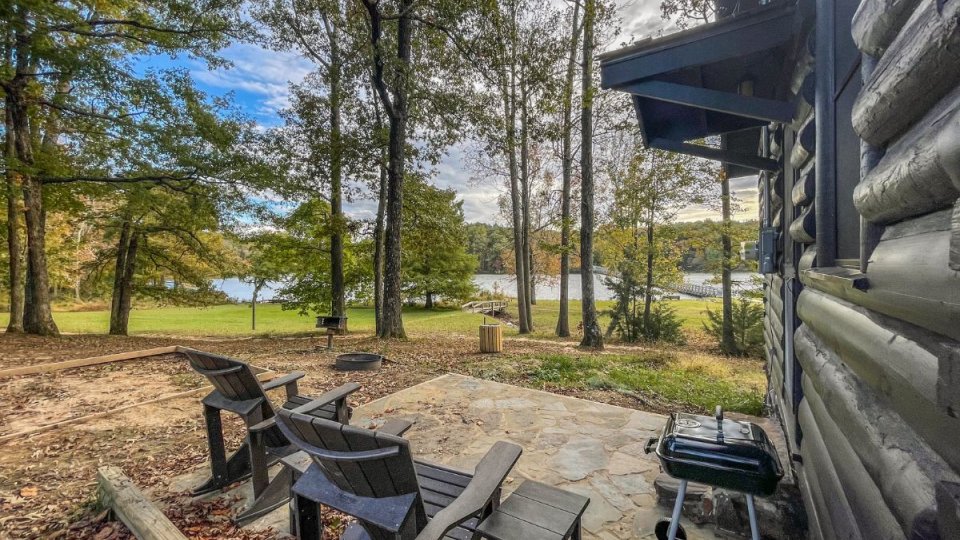 Patio with chairs beside a cabin overlooking a serene lake surrounded by trees.