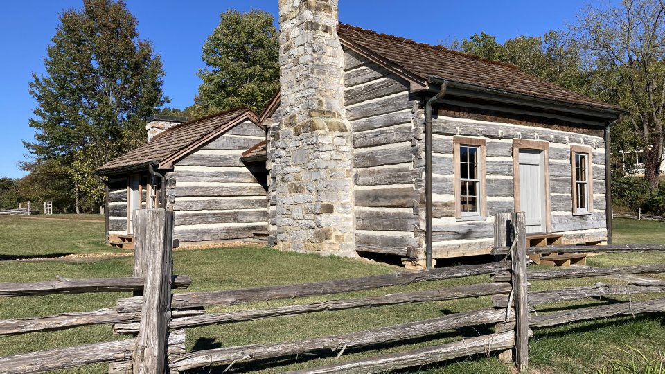A rustic wooden cabin with a stone chimney and weathered appearance sits behind a wooden fence. Surrounded by grass and trees under a clear blue sky.
