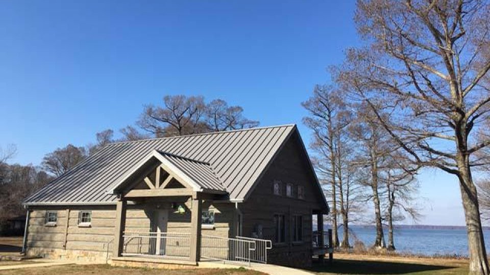 Wooden cabin by a lake with bare trees and clear sky.