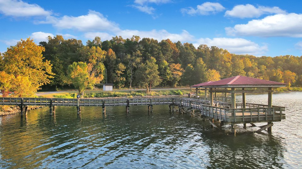 An L-Shaped fishing pier stands on the lake. Trees along the shoreline are just beginning to show their fall colors against a bright blue sky.