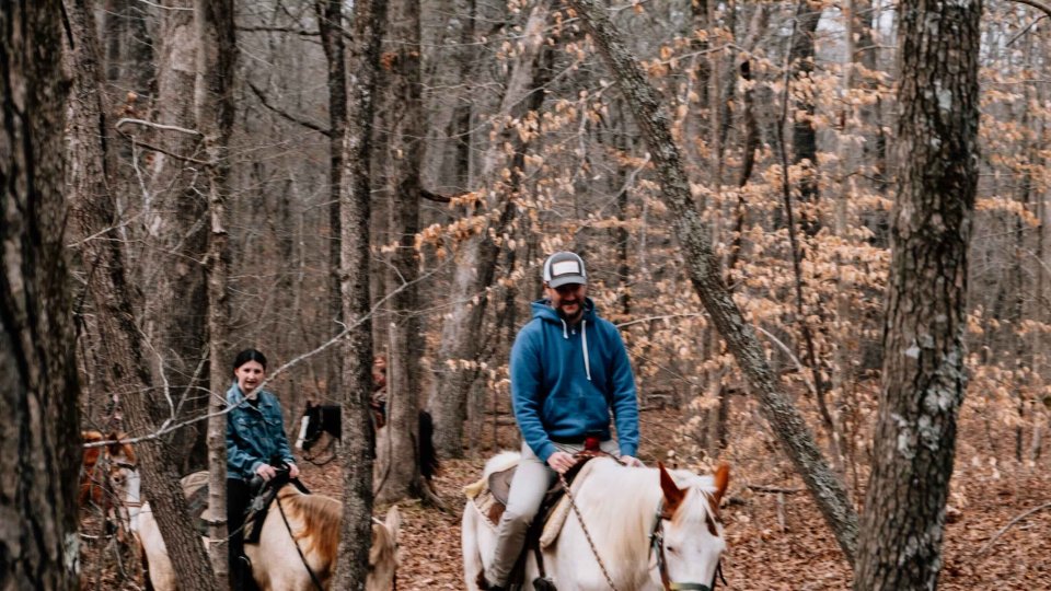 Two people horseback riding in a leafless forest.