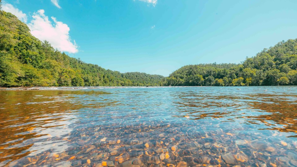 Clear river with rocky bed surrounded by lush green hills under a blue sky.