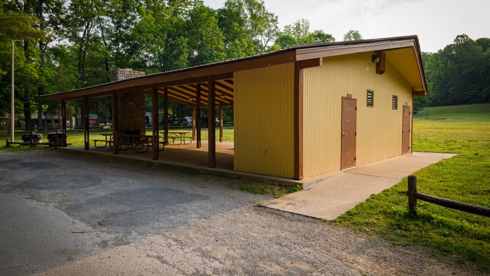A tan and brown picnic shelter stands in front of a green field lined with trees. There is a paved sidewalk on the side of the shelter and pavement in front.