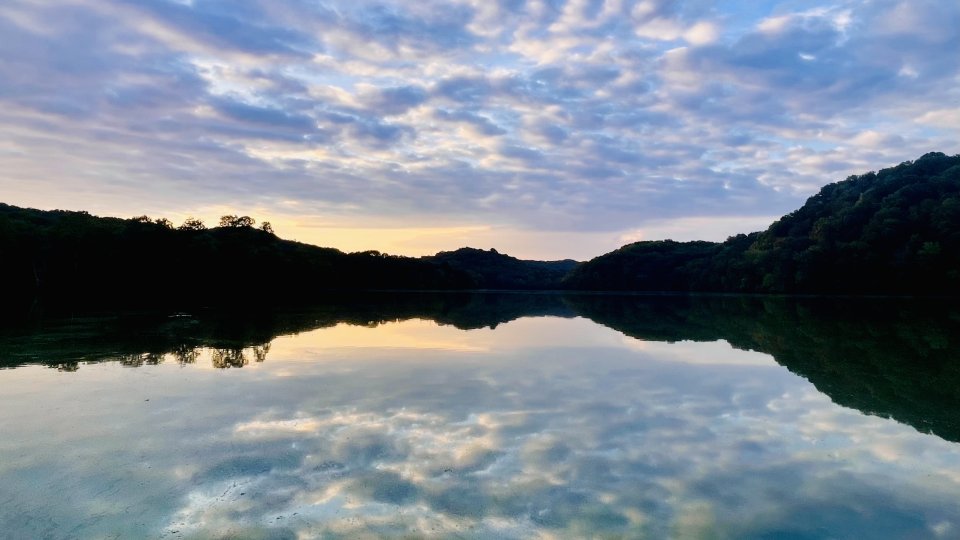 Sunset over a calm lake with reflections of clouds and tree-lined hills.