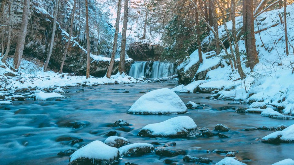 Snow-covered river with rocks and a small waterfall in a forest.
