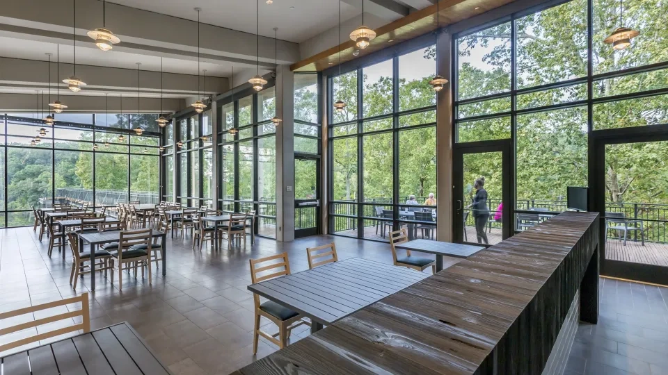 The interior of a restaurant with large floor to ceiling windows. There are tables and chairs inside and you can see outside that there is a patio with more seating options.