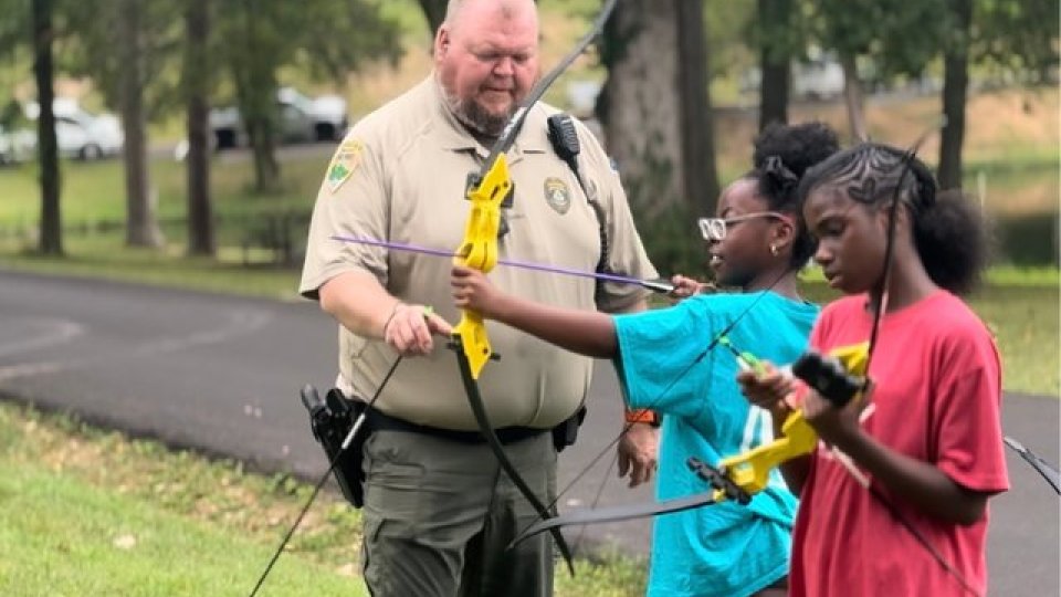 Park ranger teaching two young girls archery outdoors.
