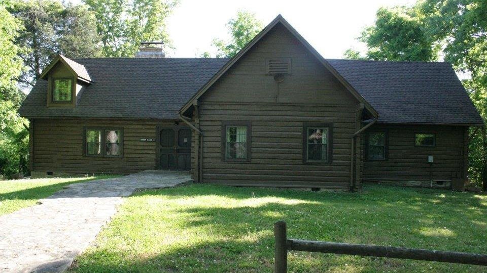 Brown wooden house with gabled roof in a green, tree-filled setting.