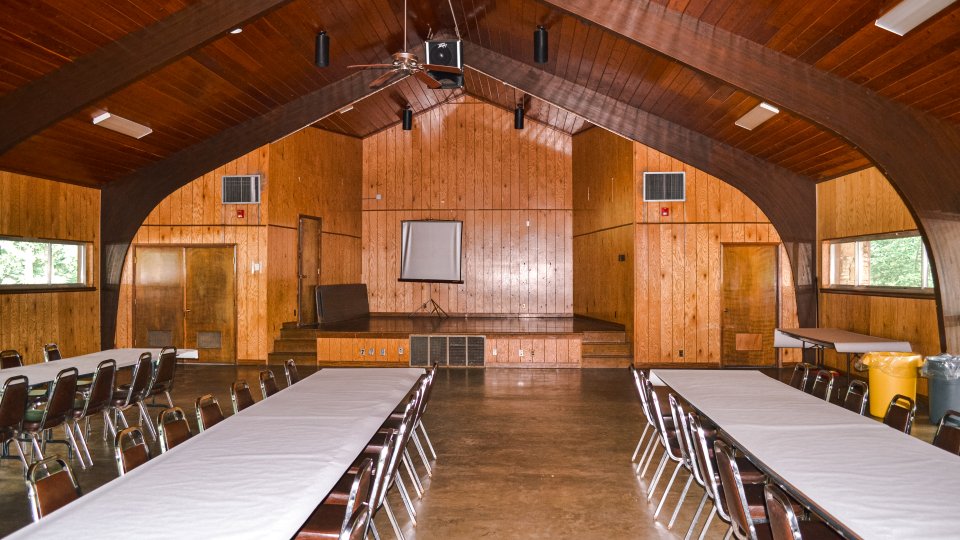Wood-paneled hall with arched ceiling, tables set in rows, and projection screen.