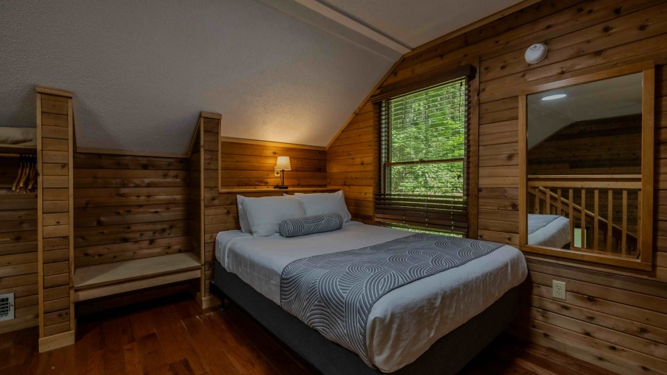 Cozy cabin bedroom with wooden walls and a large window showing greenery.