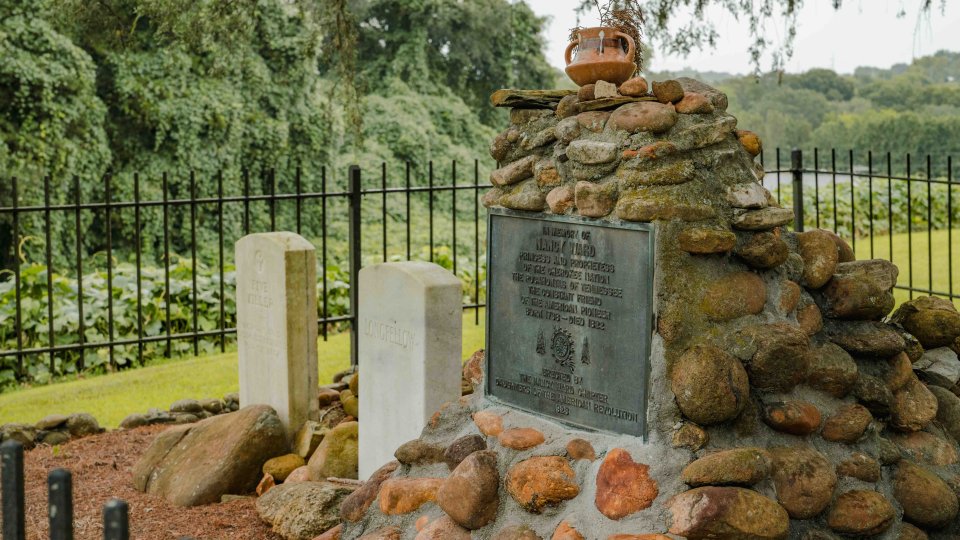 Stone memorial with plaques surrounded by greenery and fencing.
