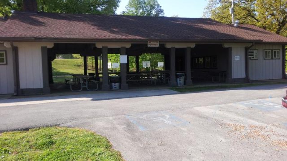 Park pavilion with picnic tables, surrounded by trees on a sunny day.