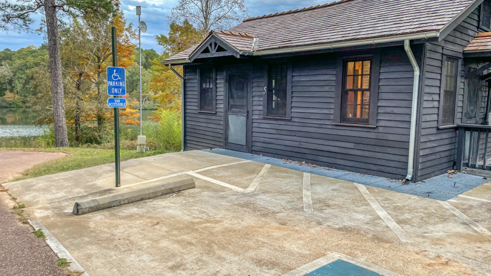 A blue ADA cabin sits behind a wheelchair accessible parking space. To the left, the lake is visible, surrounded by trees just beginning to change colors for Fall. 