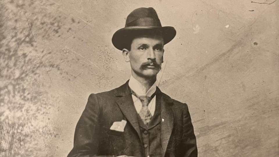 Victorian era man in suit and hat holding a cigar, standing beside a chair.