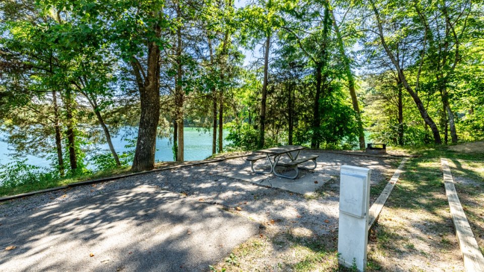 A serene RV campsite by a lake, surrounded by tall trees and greenery. A picnic table sits on gravel, with a peaceful water view nearby