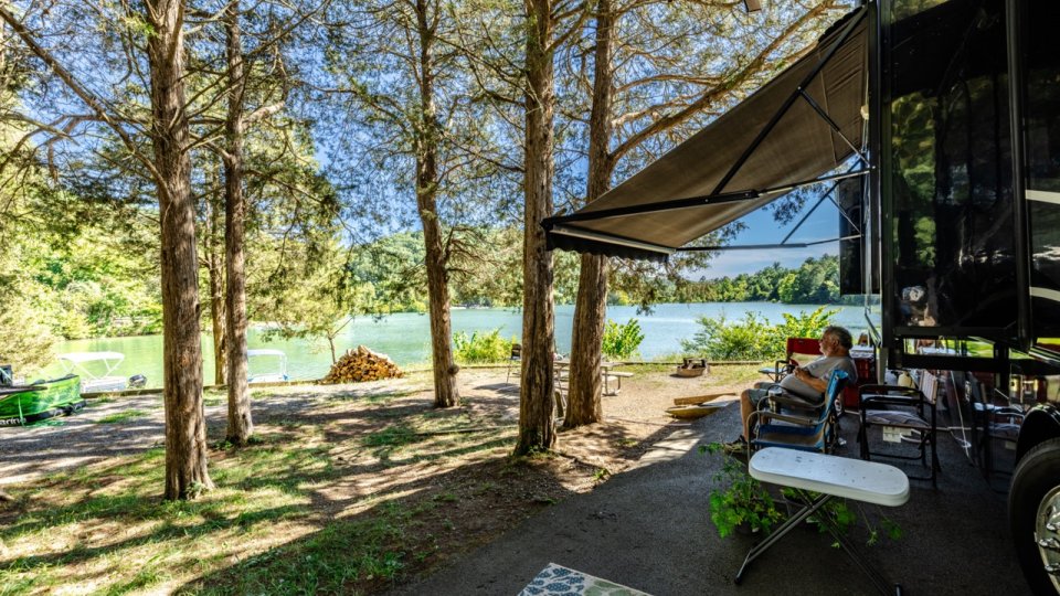 A serene lakeside scene featuring a camper under tall trees, with a person relaxing in a chair, surrounded by nature.