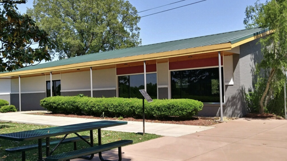 Single-story building with a green roof, trees, and a picnic table in front.