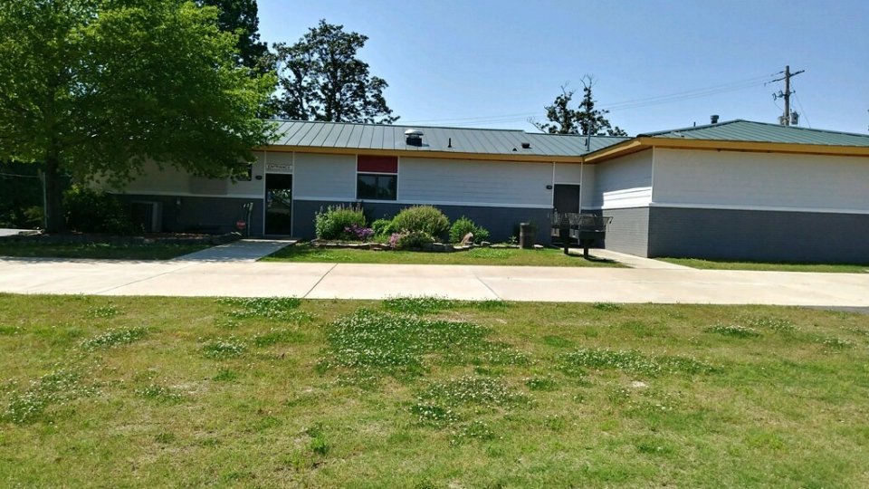 Single-story building with a gray roof, surrounded by greenery under a clear blue sky.