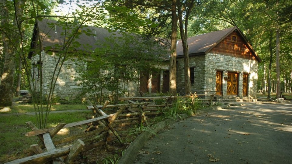 Stone house with wooden roof in a forest setting, surrounded by trees and a wooden fence.