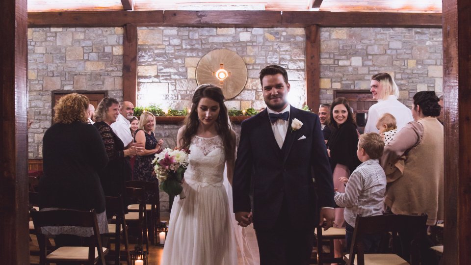 Bride and groom walking down aisle in rustic, warmly-lit venue.