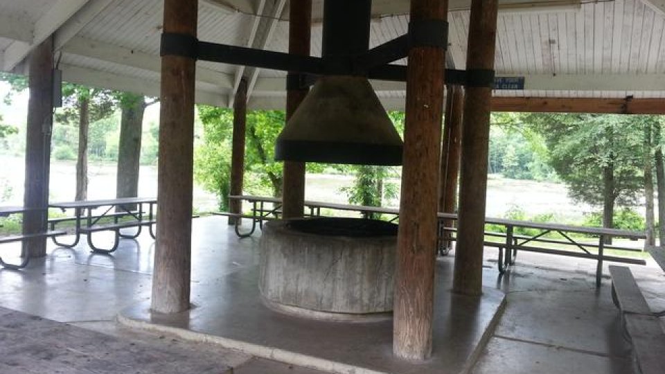 Open-air pavilion with a large bell and picnic tables, surrounded by trees.