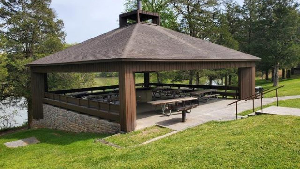 Outdoor picnic pavilion with a shingled roof by a lake, surrounded by green grass.