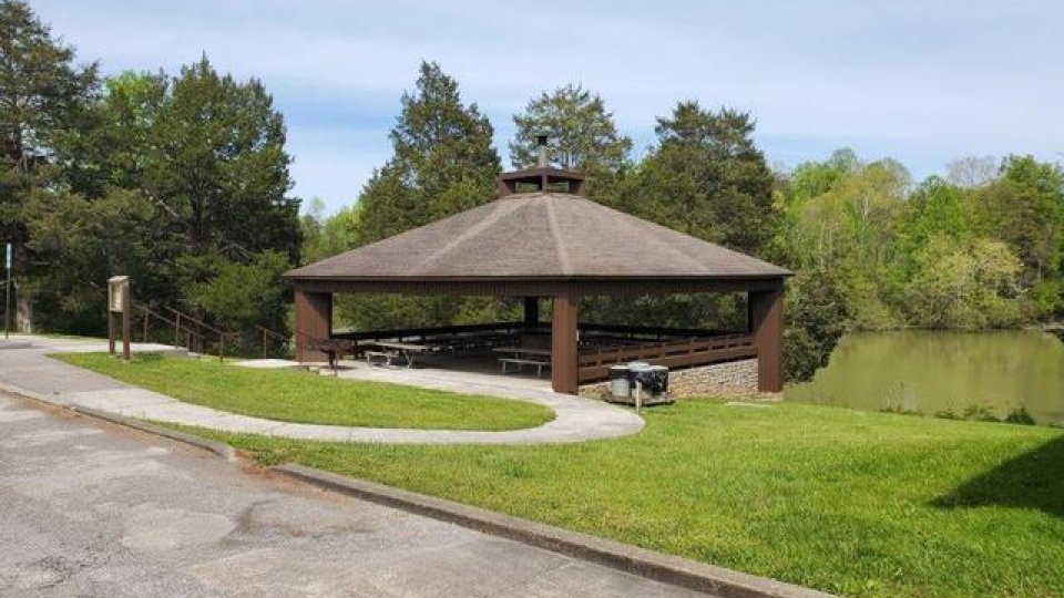 Outdoor pavilion by a lake with surrounding greenery on a sunny day.